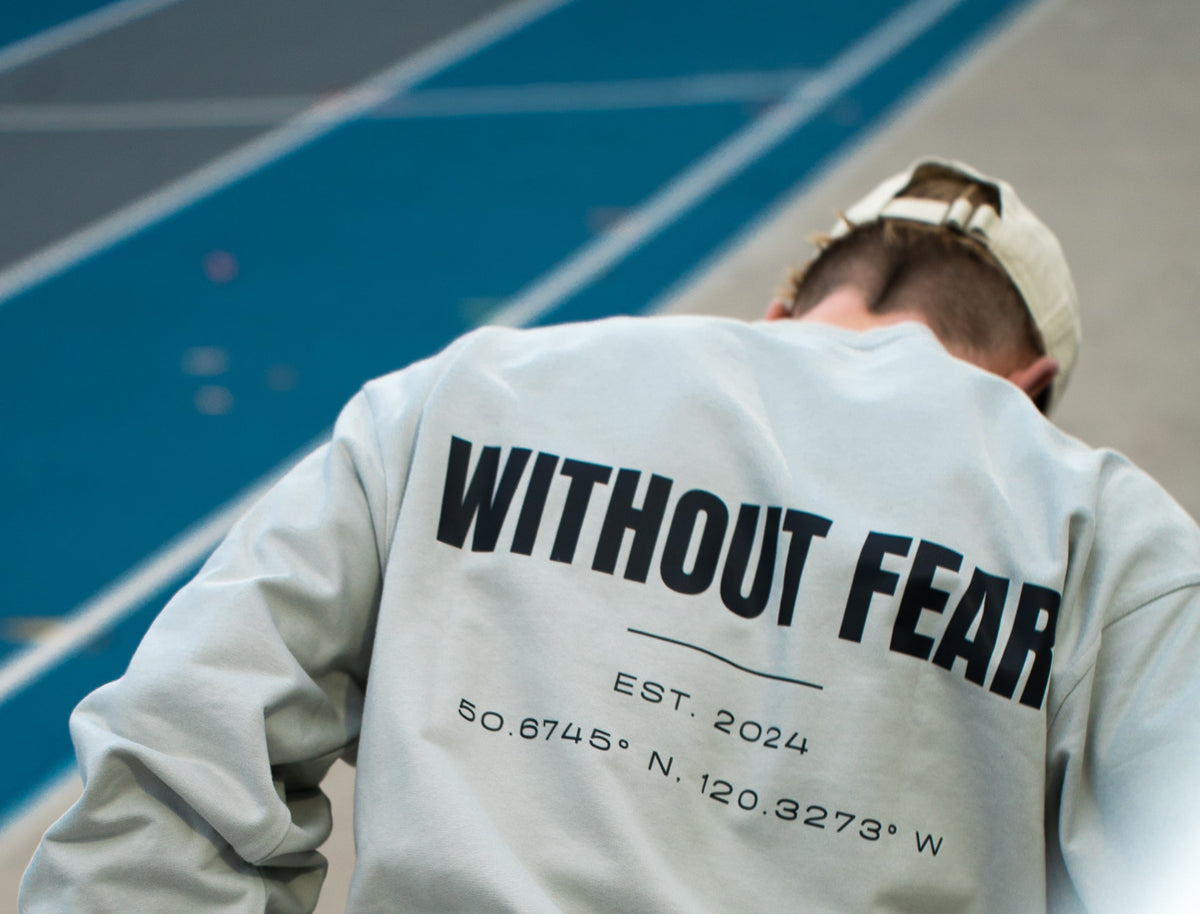 A person viewed from behind wearing a light gray sweatshirt with 'WITHOUT FEAR EST 2024' printed in black on the back, standing on a blue running track.