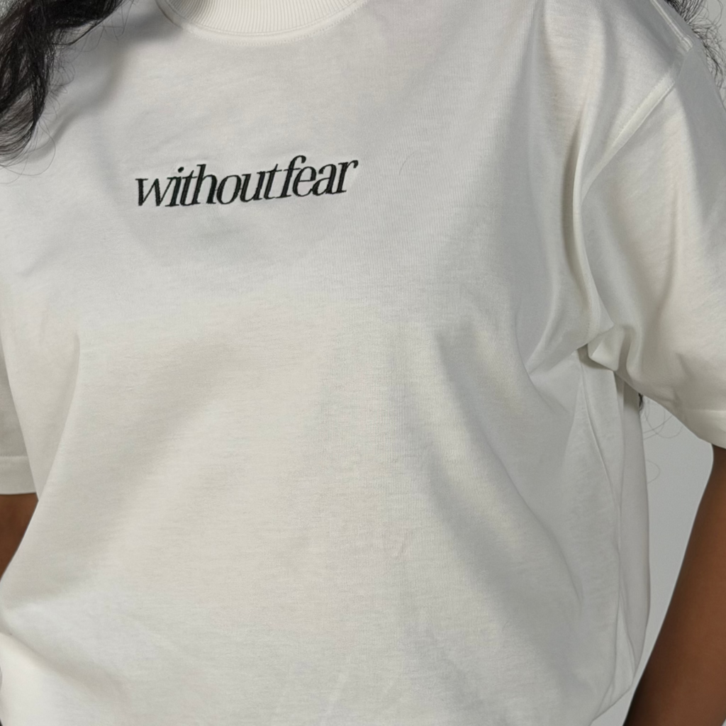 A close-up of a person wearing a white short-sleeve T-shirt with the word 'withoutfear' printed in black serif font across the chest, paired with black bottoms.