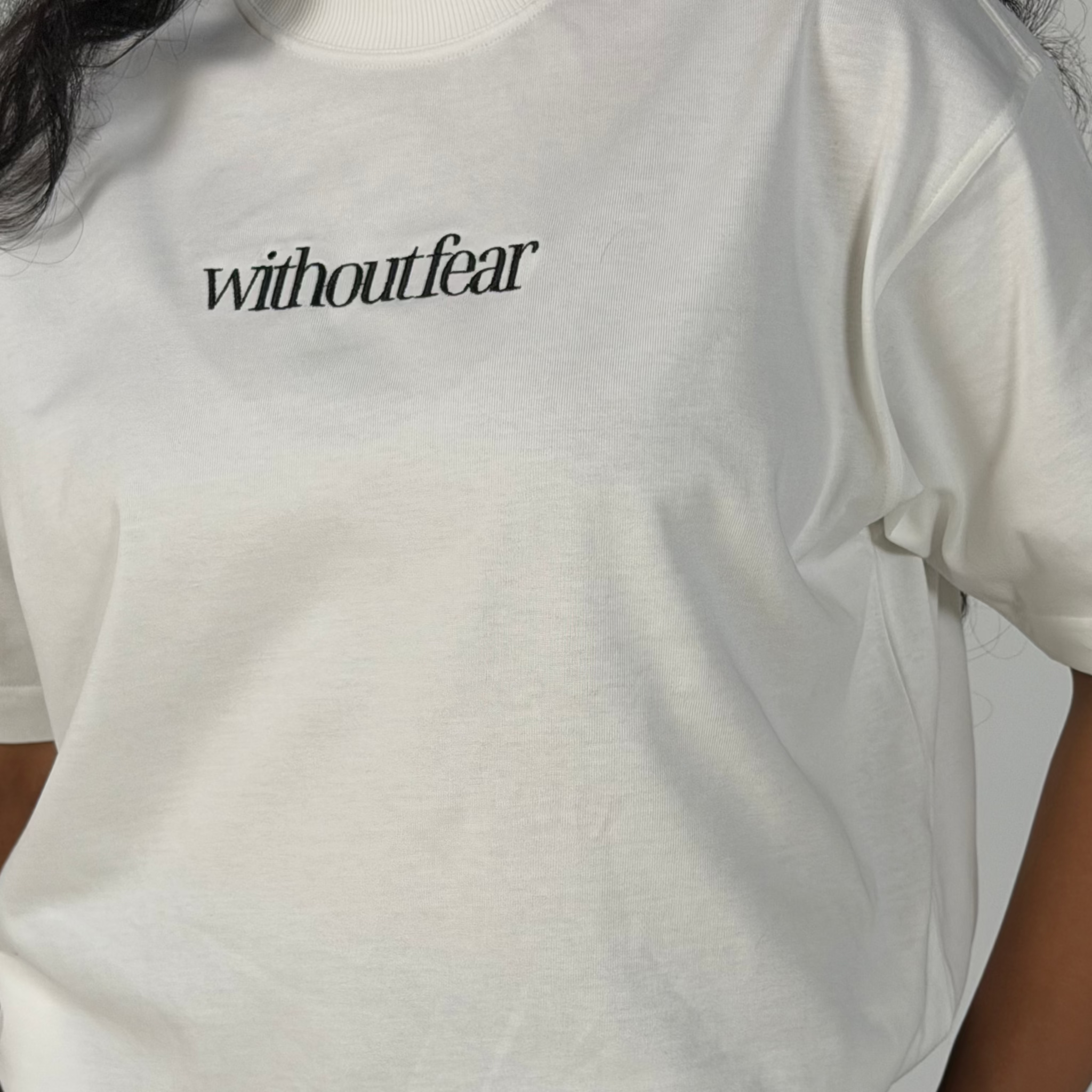 A close-up of a person wearing a white short-sleeve T-shirt with the word 'withoutfear' printed in black serif font across the chest, paired with black bottoms.